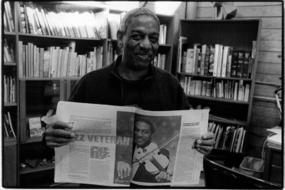 Head and shoulders portrait of Billy Bang holding a newspaper spread with a featured article about him, Woodside, CA (2005)