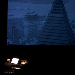 Michael Nyman, seated at piano, back to camera, with an image of city buildings projected above him, San Francisco CA., (2005)
