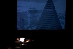 Michael Nyman, seated at piano, back to camera, with an image of city buildings projected above him, San Francisco CA., (2005)