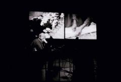 Phil Niblock seated onstage with film projections behind him, San Francisco, CA (2005)