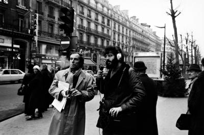 Bernard Heidsieck smokes while Charles Amirkhanian speaks into a microphone on a sidewalk in Paris, 1973