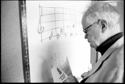 Tom Johnson, head and shoulders portrait, facing left, standing in front of a white board, holding a score, Woodside CA., vs 2 (2010)