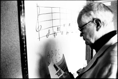 Tom Johnson, head and shoulders portrait, facing left, standing in front of a white board, holding a score, Woodside CA., (2010)