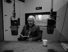Carl Rakosi, seated and reading a book, in a KPFA broadcast booth,  1971