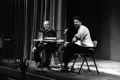 Astor Piazzolla sits at a table with Charles Amirkhanian, who is pointing something out, at the Cowell Bayfront Theater,  San Francisco, 1989