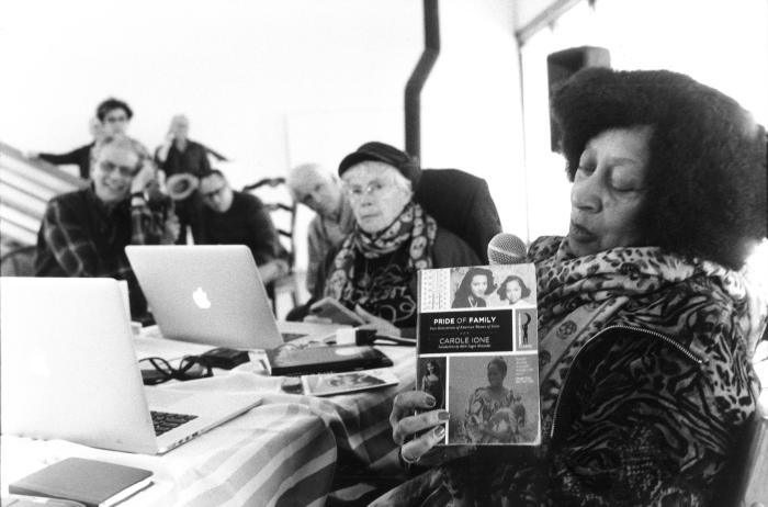 Carole Ione, holding up her book "Pride of Family" while Pauline Oliveros looks on in the background, Woodside CA (2015)