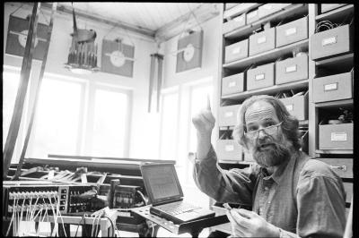 Trimpin, head and shoulders portrait, seated in his studio, facing slightly forward and pointing up, Seattle WA., (1998)