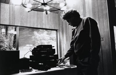 Philip Glass, three quarter length portrait, standing, adjusting stereo equipment, Woodside CA, (1993)