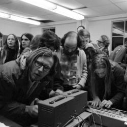 Two men adjust the setting on synthesizers while Tom Zahuranec and others look on, at a KPFA Radio Event, Mills College, Oakland CA, 1971