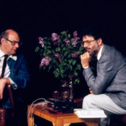 A three quarter length portrait of Mauricio Kagel seated onstage with Charles Amirkhanian during Speaking of Music at the Exploratorium, San Francisco (1988)