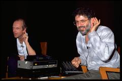 John Oswald and Charles Amirkhanian seated onstage, "listening" with hands to ears at the Exploratorium, San Francisco (1990)
