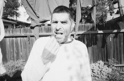 Anthony Gnazzo, head and shoulders portrait, standing in a backyard, facing forward, eating a fig, Oakland CA., (1982)