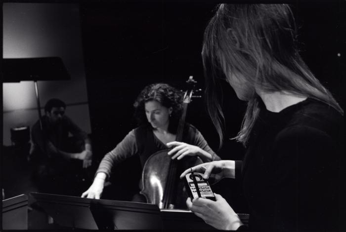 Gianna Abondolo, sitting and playing the cello, with a seated man on left and Catherine Lamb on right watching, (2009)