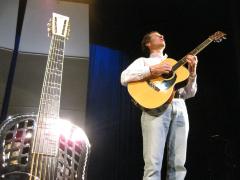 John Schneider, standing and playing the guitar, with his re-fretted steel guitar standing on the left, during OM 14, ver. 1.