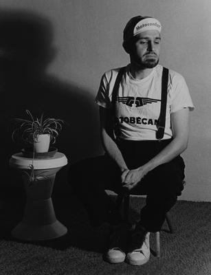 Portrait of Anthony Gnazzo sitting on a stool next to a plant (undated)