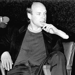 Brian Eno, seated onstage with leg over chair arm during Speaking of Music at the Exploratorium, 1988