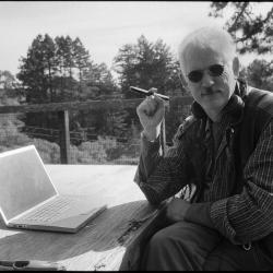Half length portrait of Kyle Gann, seated outdoors with a laptop and cigar, Woodside CA (2011)