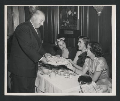 Portrait of George Mardikian offering lavash to Anahid and Maro Ajemian, seated with Nazenig Mardikian at Omar Khayyam's Restaurant in San Francisco (ca.1950s)