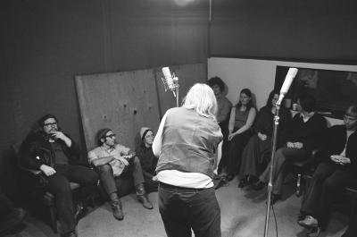 A group of people sitting in the KPFA studio while staff set up microphones (1972)