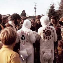 Two people in bunny costumes, as seen from behind, surrounded by a crowd of people, in a park like setting