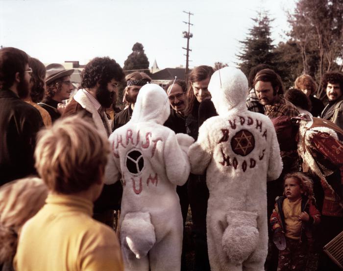 Two people in bunny costumes, as seen from behind, surrounded by a crowd of people, in a park like setting